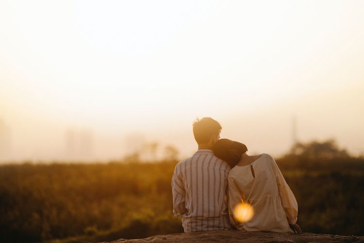 Man and woman sitting facing away, her head on his shoulder, looking at grass field and setting sun